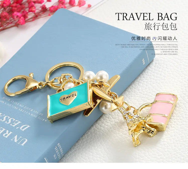 Llavero de viaje en tono dorado con un colgante de bolsa de viaje de esmalte turquesa, detalles de perlas y una Torre Eiffel esmaltada en rosa con adornos de cristal.