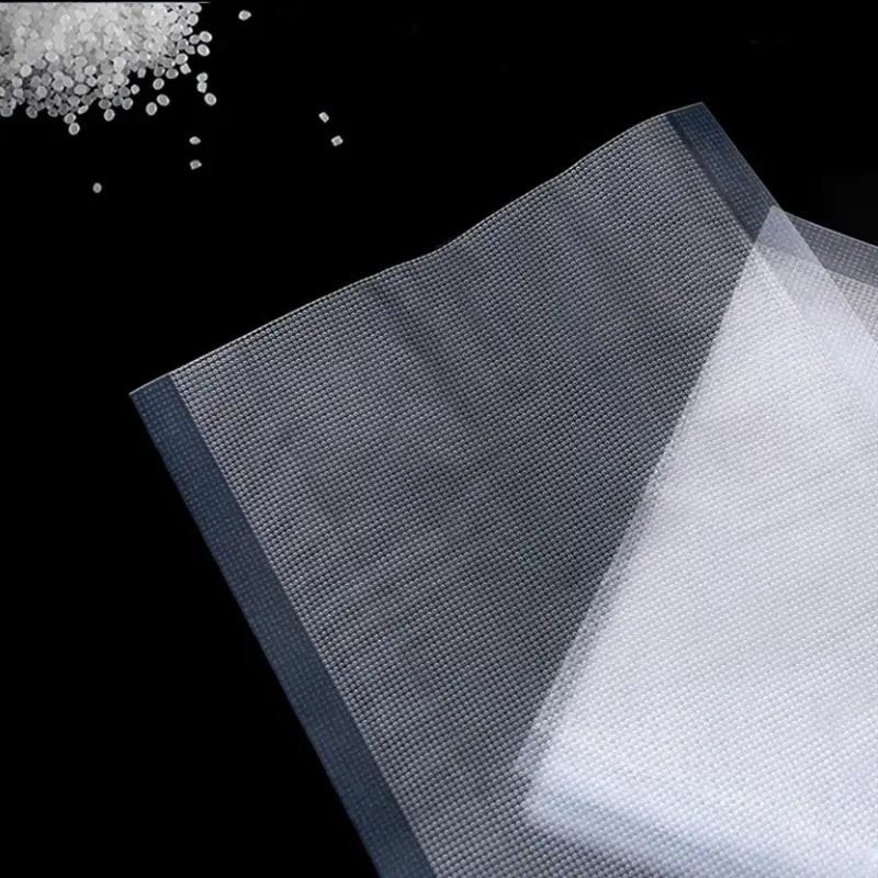 Strukturierte Vakuumbeutel 12x20 cm mit transparentem Mesh-Gewebe mit dezentem Rastermuster und verstärkten Kanten, dargestellt zusammen mit weißen Kunststoffgranulaten zur Lebensmittelversiegelung