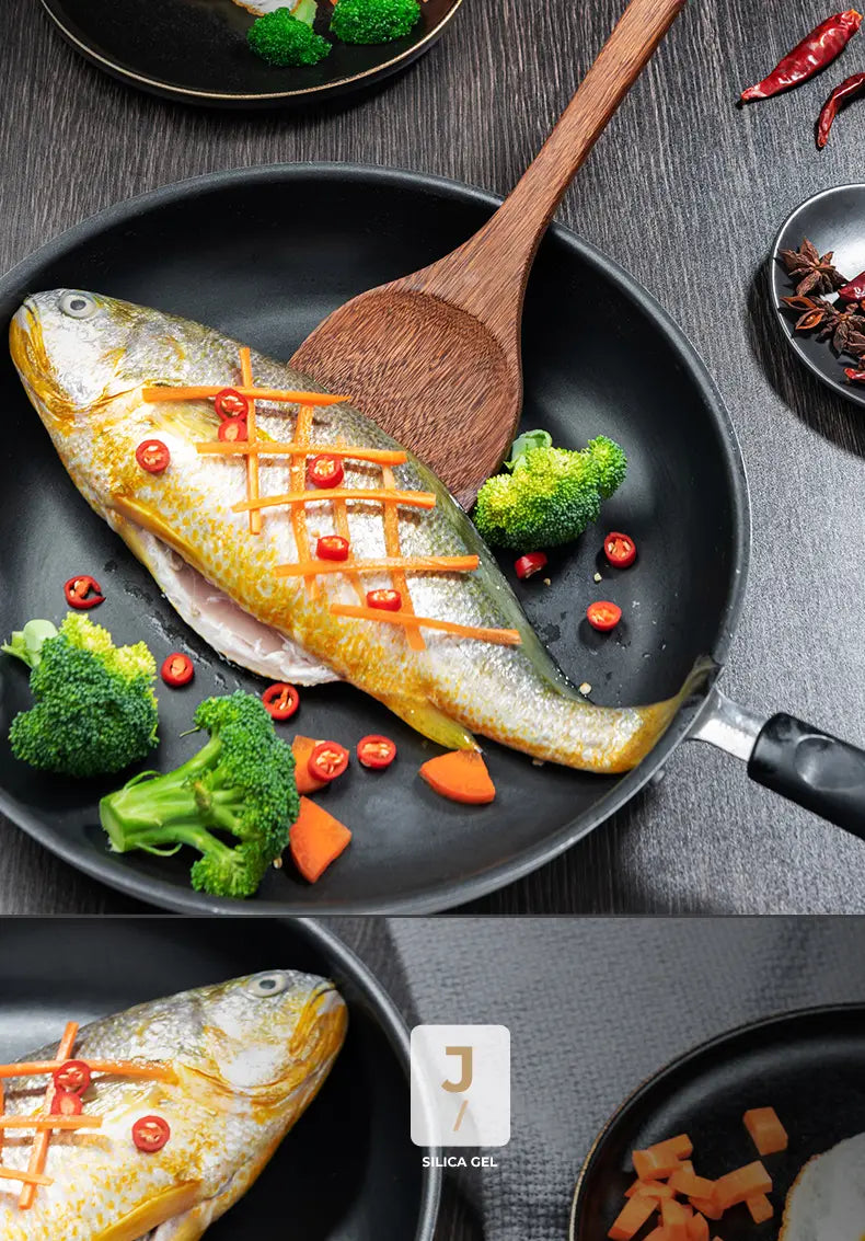 a golden brown whole fish garnished with orange carrot strips and red chili slices rests in a black non-stick frying pan suitable for use with a wooden kitchen utensil set non-stick cookware.