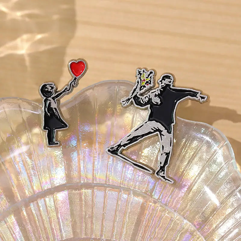 Épingle émaillée « Banksy » de type broche, représentant un duo d’épingle émaillées à motifs emblématiques noir et blanc rehaussés de couleurs vives, illustrant un enfant tenant un ballon en forme de cœur rouge et une silhouette lançant des fleurs.