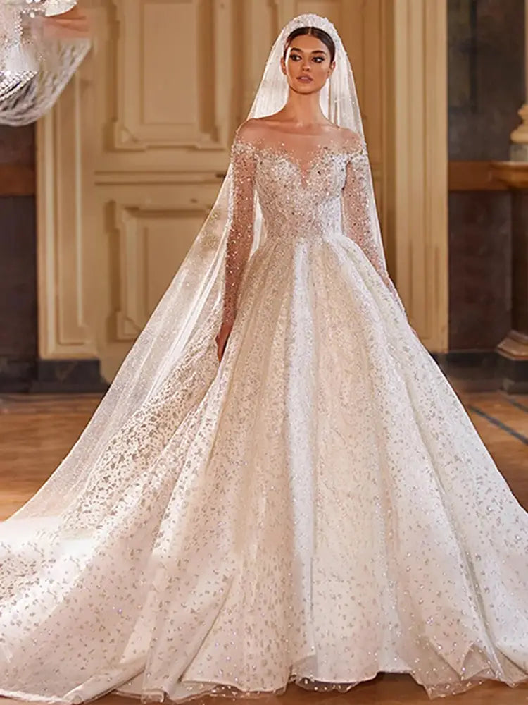 Une robe de mariée en style boule ivoire étincelante, agrémentée de perles, avec des manches longues et des détails en dentelle, ainsi qu'une traîne fluide.