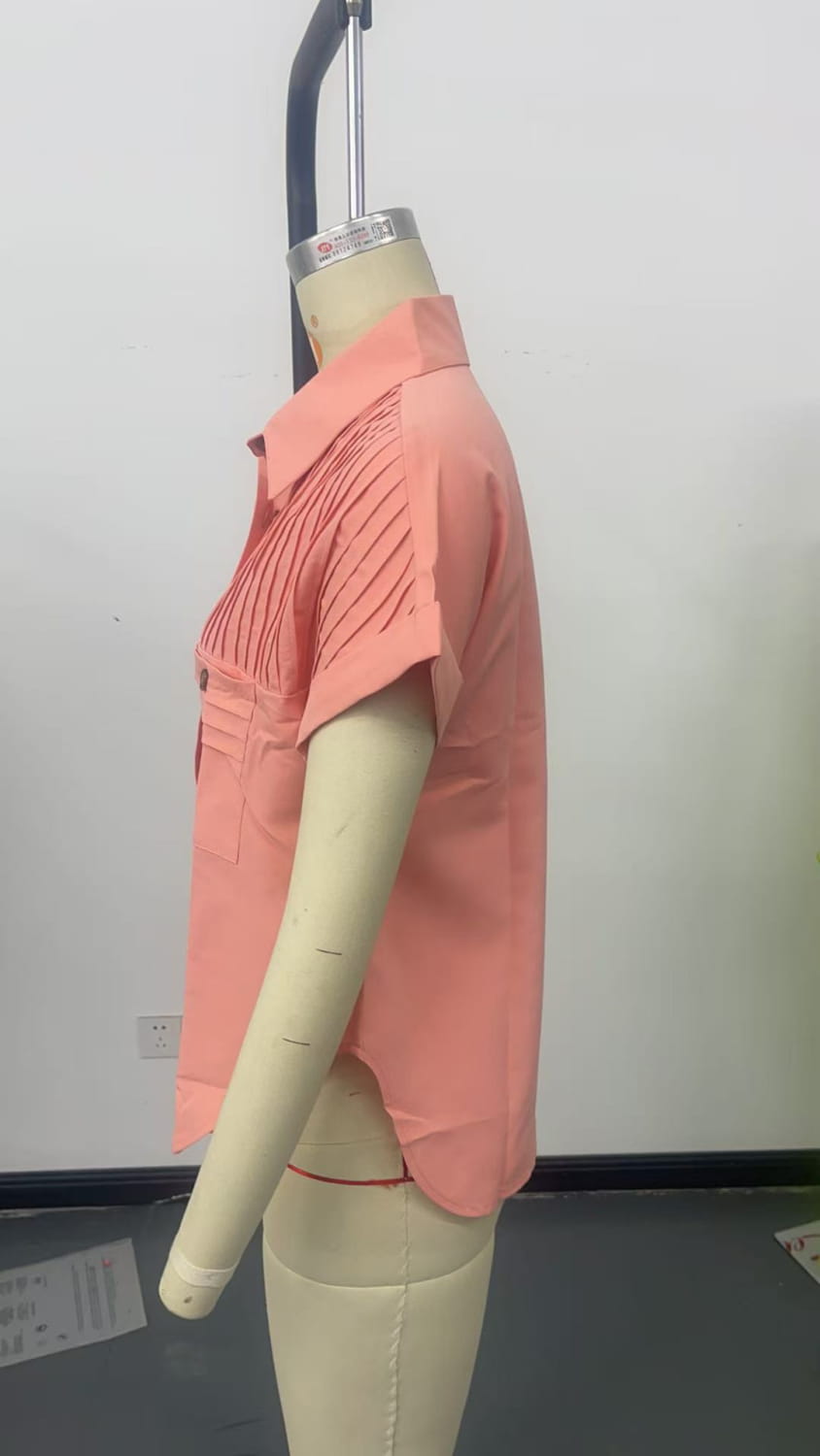 A coral-colored short sleeve button up blouse for women featuring pleated detailing on the chest and a buttoned front with short sleeve length.
