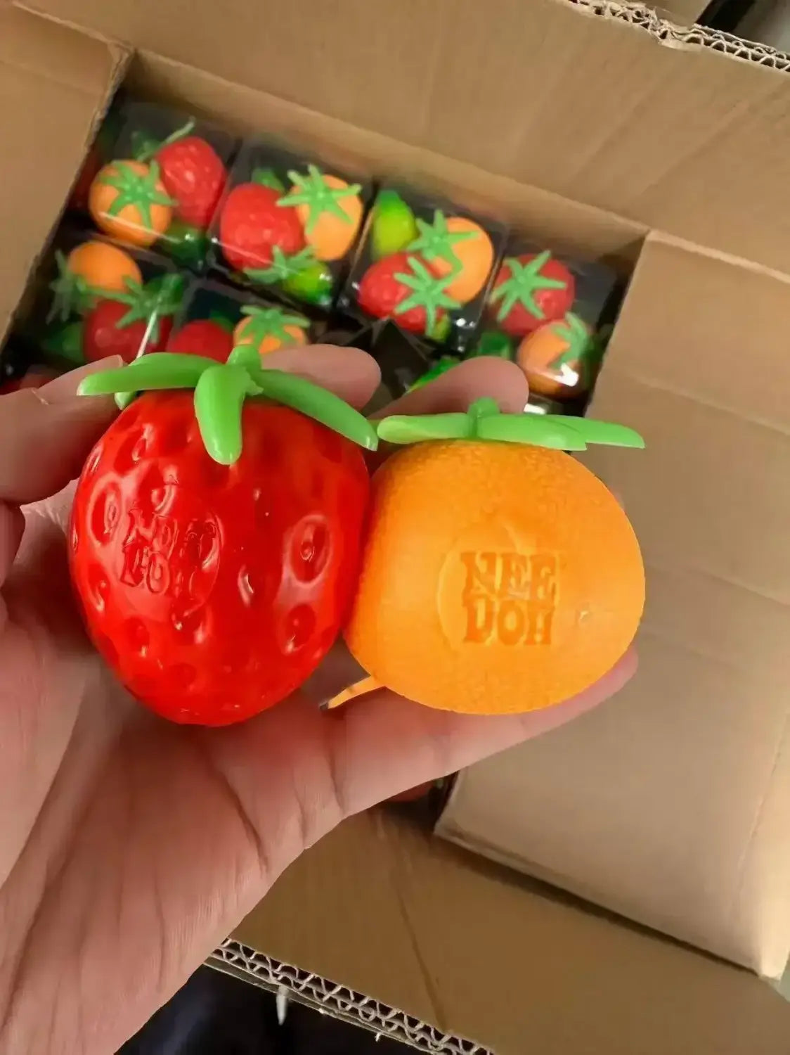 Ein rotes und orangefarbenes, quetschbares Spielzeug in Form einer Erdbeere und einer Orange mit grünen Stielen, aus der Frontansicht im Erdbeere-Orange-Banane-Stressabbau-Set.