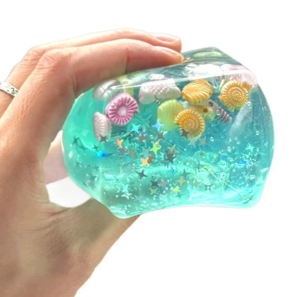 Un cube sensoriel à effet anti-stress pour l’océan en turquoise translucide, rempli de charmes de coquillages colorés et de confettis scintillants en forme d’étoiles, conçu comme un cadeau attentionné.
