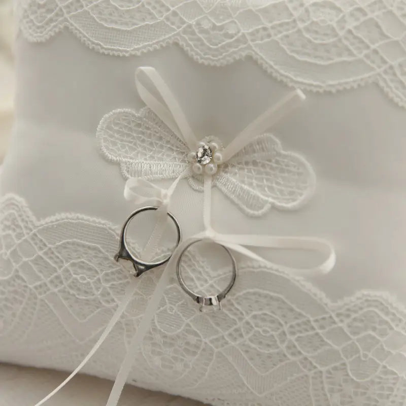 Almohada blanca para anillos de boda de encaje con aplicación de mariposa y almohadilla portadora de arras con dos anillos que descansan sobre ella.