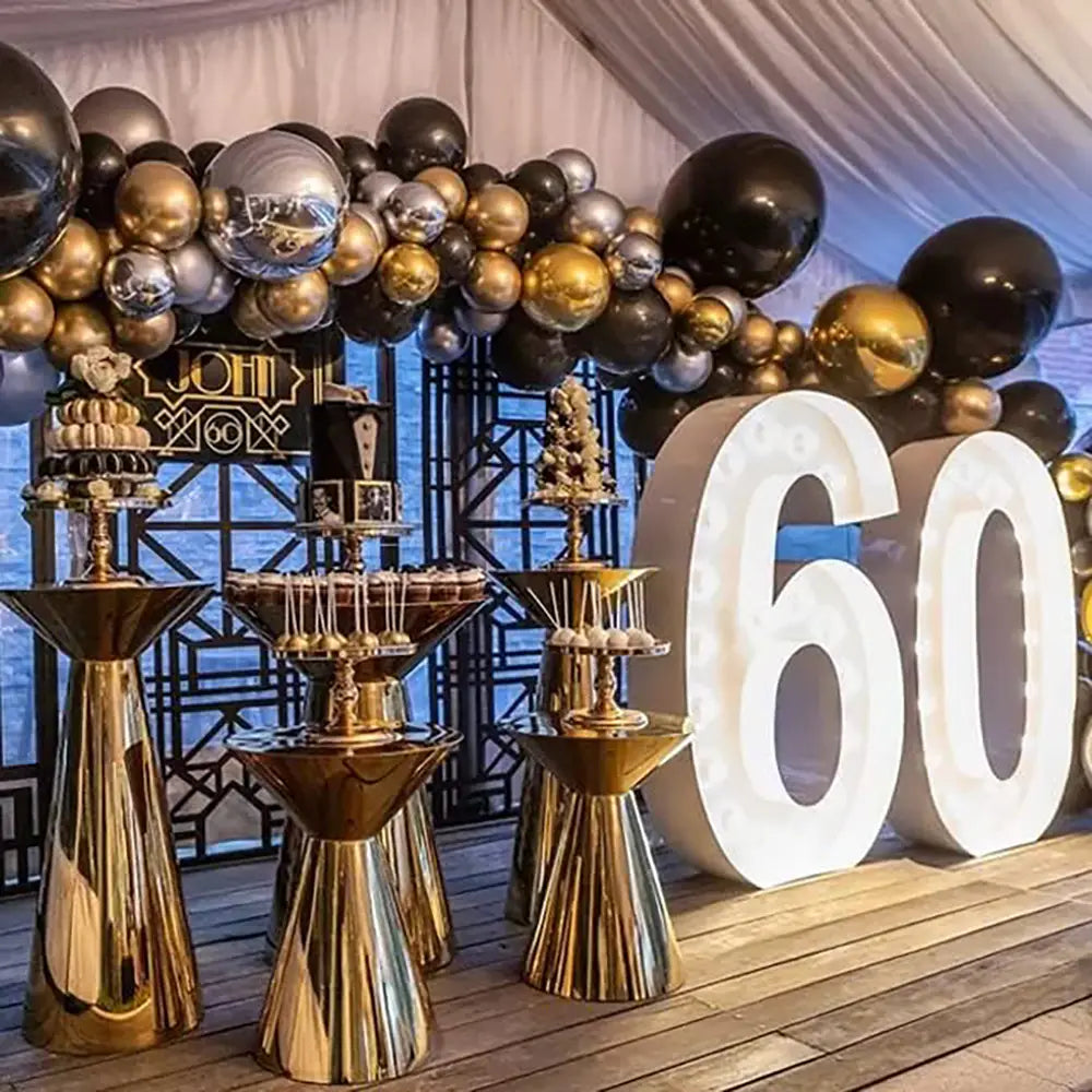 Glänzende schwarze, goldene und silberne Ballons, in einem Bogen über einer festlichen Dessertpräsentation angeordnet; zeigt das schwarze und goldene Ballonbogen-Set für Partys.