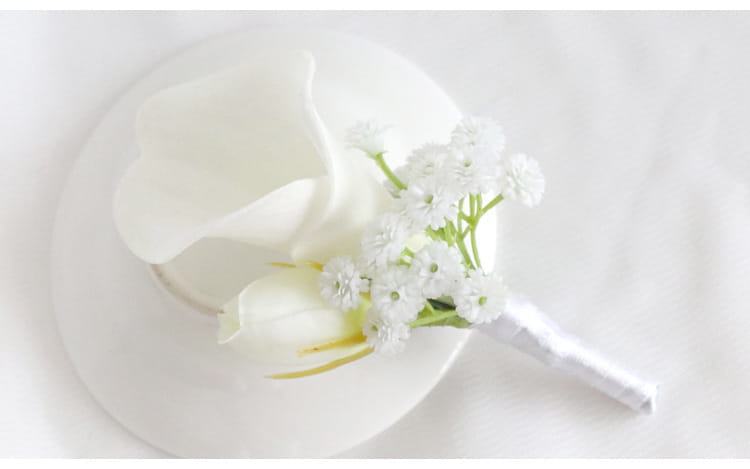 大きなカラーリリーと、控えめな緑の茎でアクセントを施したベビーズブレスの房をあしらった、繊細な白い人工カラーリリーのウエディング・ボタンニエール。ボタンニエールおよびリスト・コサージュのセットに含まれます。