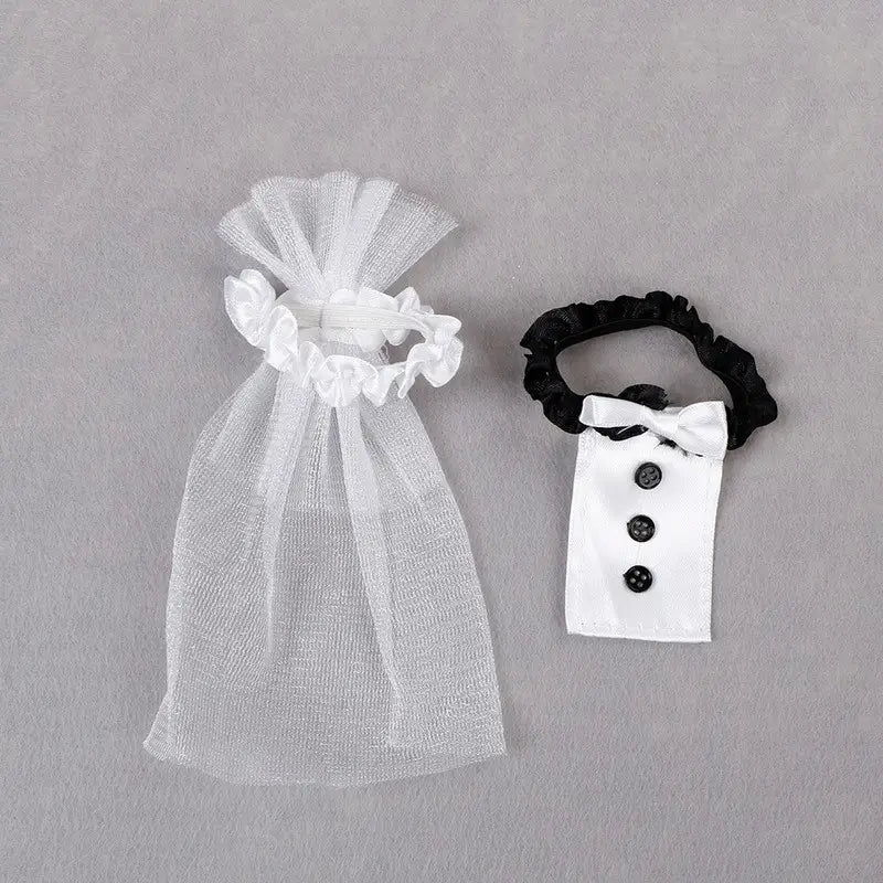 Bolsa de regalo de organza blanca semitransparente con parte superior fruncida tipo cordón y un coletero elástico negro a juego, adornada con un moño de corbatín blanco en miniatura y botones negros para el juego de cubiertas de copas de vino para la novia y el novio.