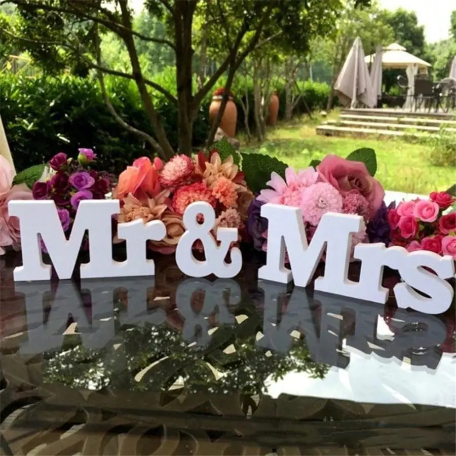 Weißes hölzernes Hochzeitstischschild mit den Buchstaben „Mr“ und „Mrs“, als Dekoration auf einer reflektierenden Oberfläche ausgestellt, umgeben von farbenfrohen Blumen um die Holzbuchstaben