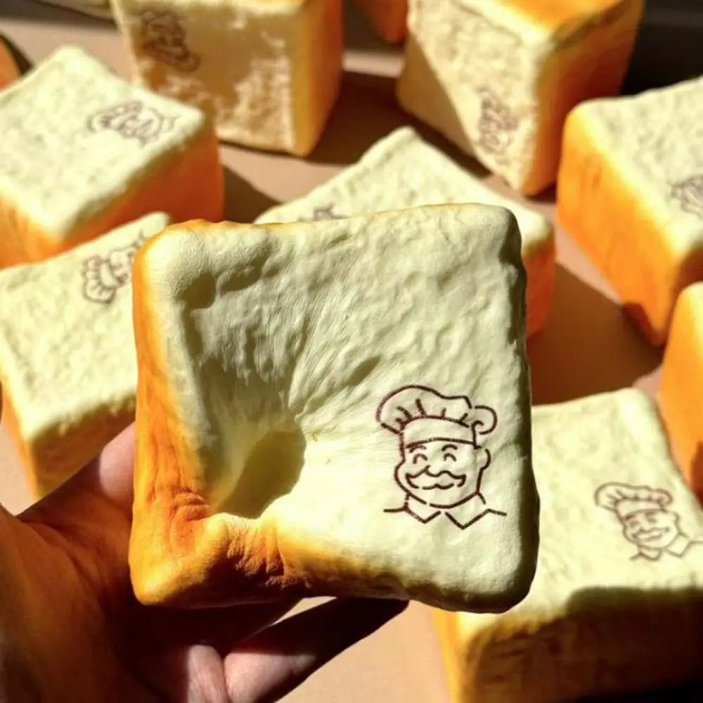 Soft squishy toy shaped like bread with a golden-brown crust and pale interior featuring an embossed cartoon chef face viewed from the front, designed as a sensory fidget toy for a universal age range.