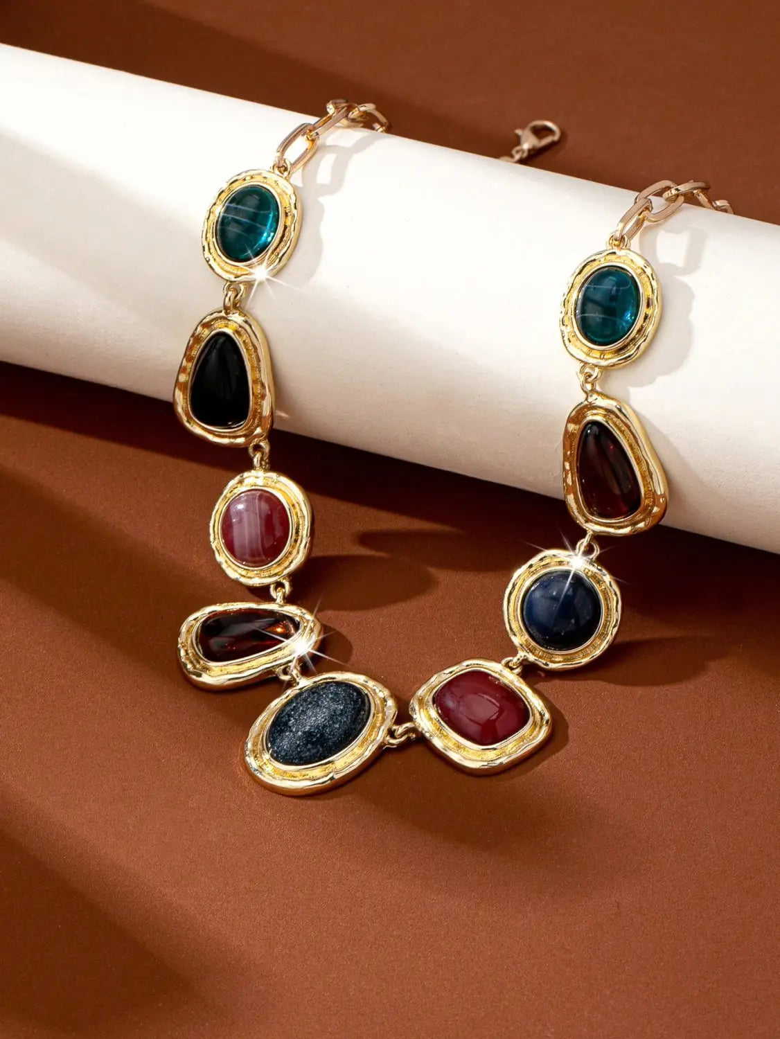 Collier pendentif en pierre précieuse géométrique à finition dorée pour femmes, présentant des pierres précieuses en forme ovale et de goutte dans des tons vibrants de bleu-vert, de noir, de rouge profond et de bleu marine.