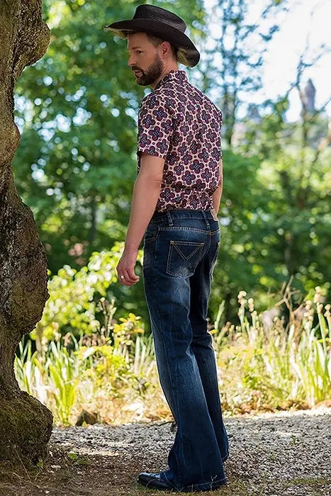 Ein Mann trägt Herren ausgestellte Jeans im Bell-Bottom-Stil aus dunkelblauem Denim, kombiniert mit einem schwarzen Cowboyhut und einem kurzärmeligen Hemd mit lila-weißem floralen Musterdruck.