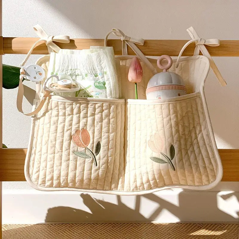 クマの刺繍が施されたクリーム色のキルティング製ベビーカー用オーガナイザー袋で、刺繍のチューリップのデザインと、赤ちゃんの着替えに必要な小物を収納するための柔らかな布製の紐が特徴です。