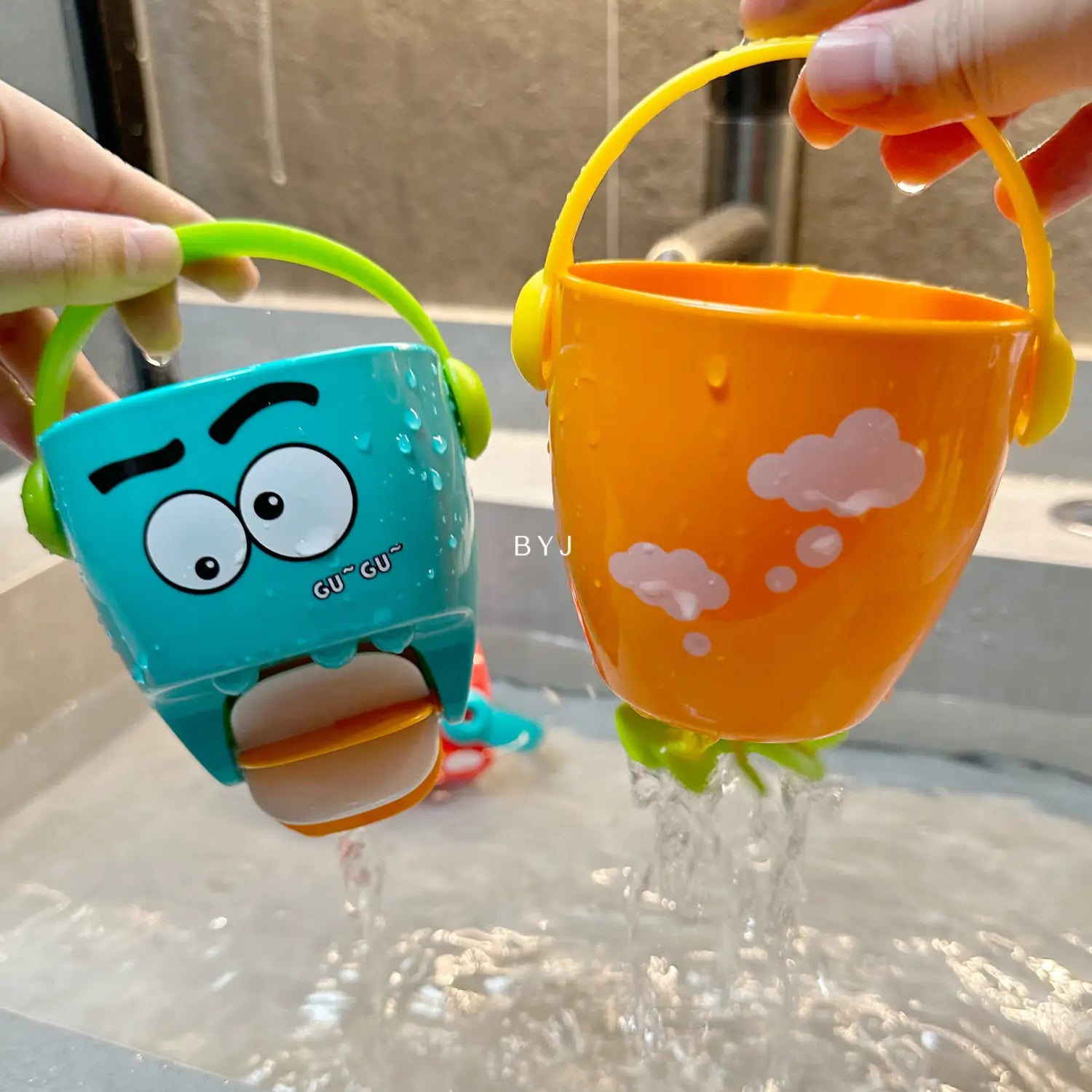 Zwei farbenfrohe Kunststoff-Badewannen-Eimer für Babys mit Wassersprühfunktion als Spielset: ein türkisfarbener Eimer mit Cartoon-Gesicht und grünem Griff sowie ein orangefarbener Eimer mit rosa Wolkenaufklebern und gelben Griffen.