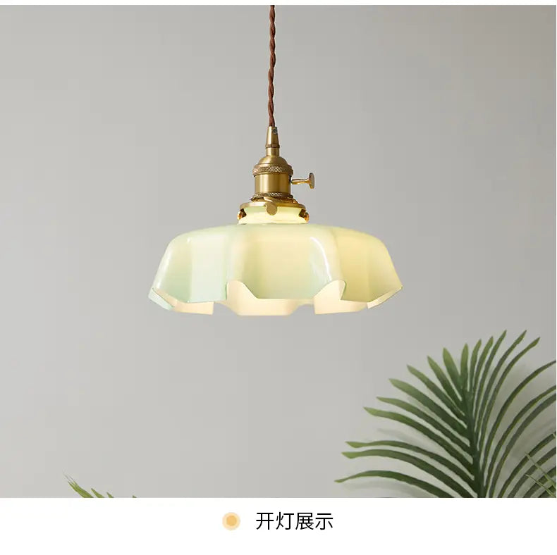 Un luminaire suspendu de style vintage avec une tête de lampe en cuivre à finition laiton et un abat-jour à jupe florale en verre blanc cassé aux bords froncés, vu depuis l’avant.