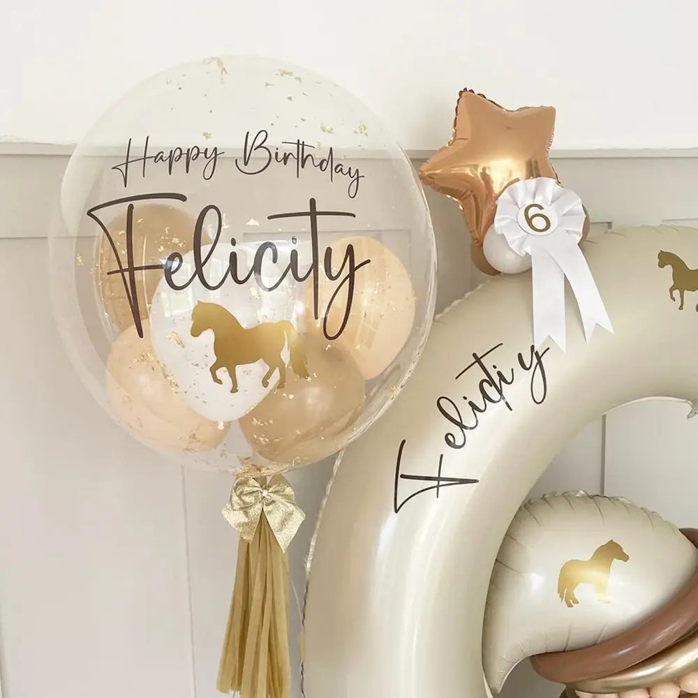 Individueller Baby-Party-Ballonaufkleber auf einem transparenten 36-Zoll-Bobo-Ballon, mit einer goldfarbenen Folien-Pferdeabbildung und „Happy Birthday Felicity“ in schwarzer geschwungener Handschrift, gefüllt mit beigem Konfetti und gebunden mit einem goldfarbenen Quastenanhänger.