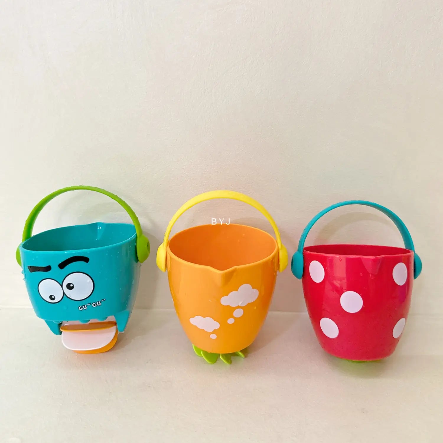 Spielset für Baby-Badewannen-Eimer mit Wassersprühfunktion, mit drei farbenfrohen Kunststoff-Strandeimern: ein türkisfarbener Eimer mit Cartoon-Augen und Tragegriff, ein orangefarbener Eimer mit weißen Wolkenmustern und grünen Füßen sowie ein rot gepunkteter Eimer mit passendem tealfarbenen Griff.