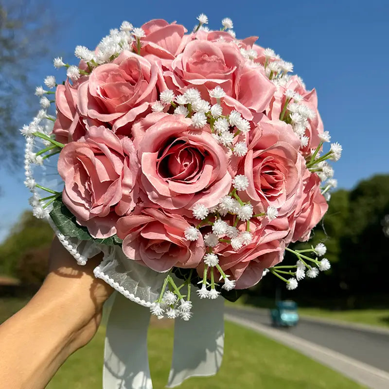A delicate pink artificial rose bouquet accented with white baby’s breath and tied with a lace-edged ribbon for a white silk rose wedding bouquet with lace and pearls.