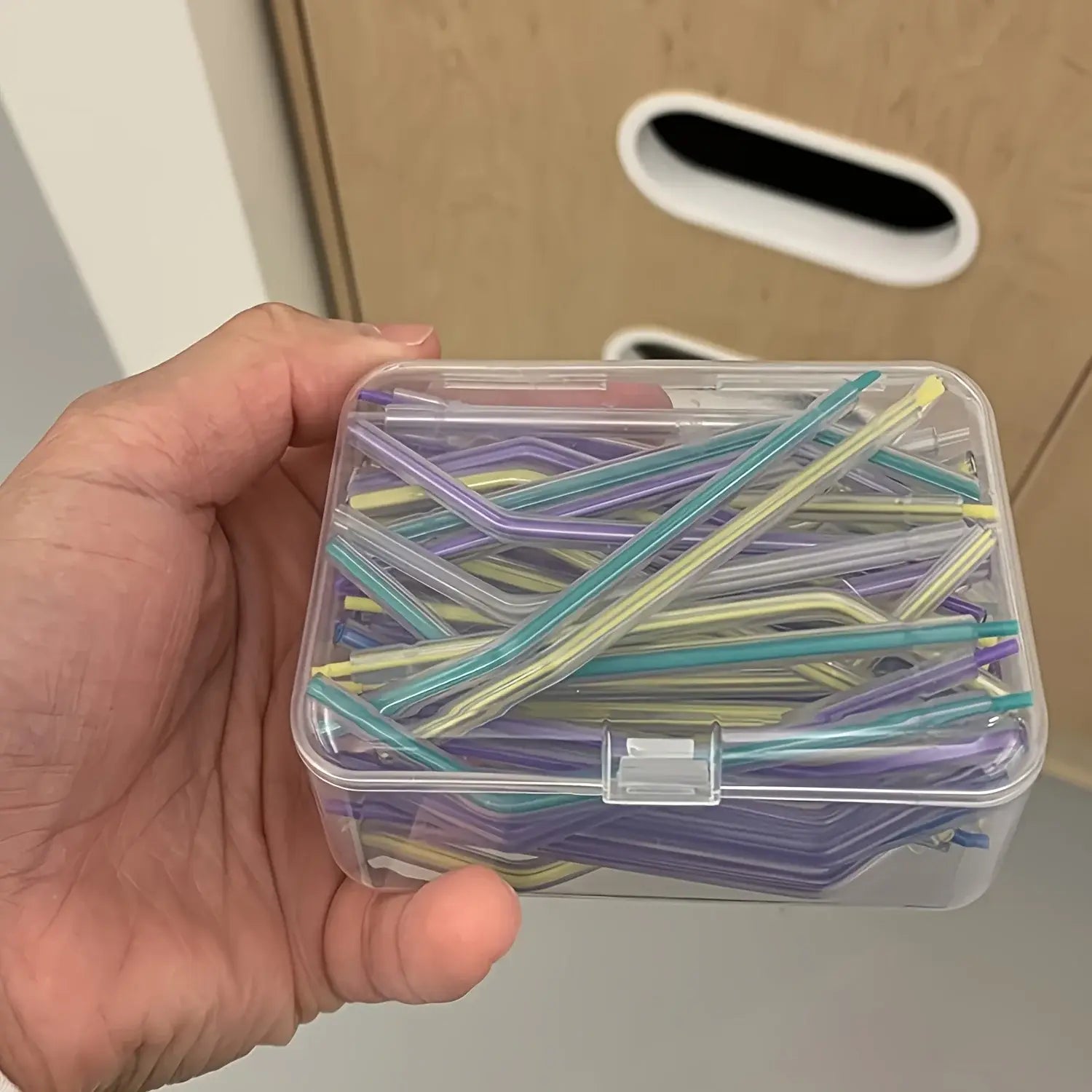 Une boîte de rangement en plastique transparent contient un ensemble de pailles en verre translucide de couleurs variées, dans des tons de violet, de turquoise et de jaune, pour organiser des travaux manuels et des strass.