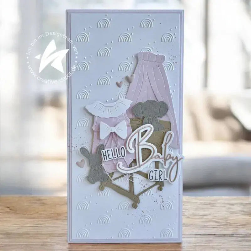 Eine zarte Lavendel-Grußkarte für ein Baby-Mädchen mit übereinanderliegenden Papier-Ausschnitten von Kleid, Mützchen und Kinderwagen mit metallisch goldenen Akzenten, hergestellt mit Oster-Ei-Hasen-Babyschneidvorlagen