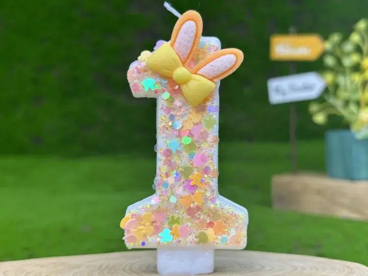 A glitter bunny ear number birthday candle featuring a glittery number one adorned with a yellow bow and orange bunny ears.