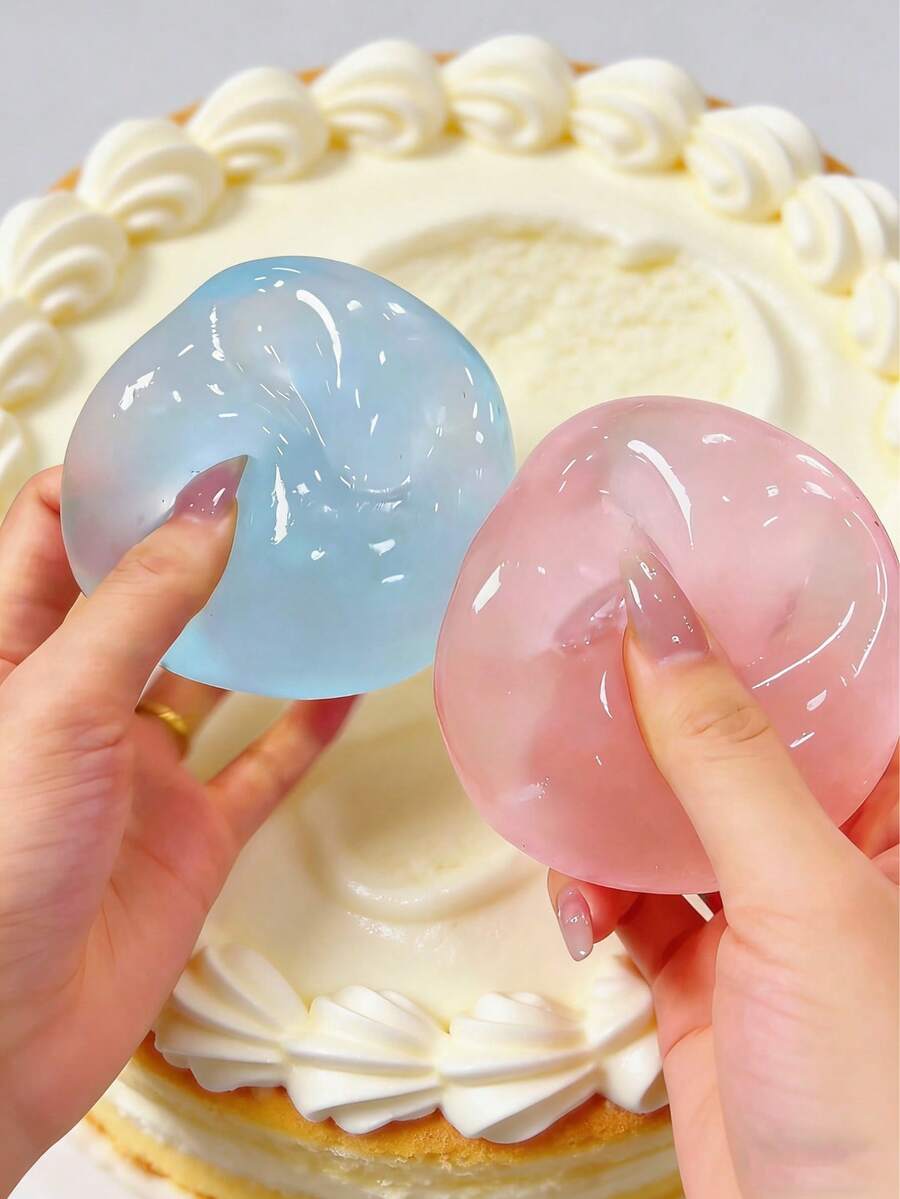 Deux balles anti-stress translucides et souples, bleu clair et rose, tenues dans des mains soignées, contre un arrière-plan de gâteau glacé blanc, jouet de compression « Blue Coconut Oil Stress Ball ».