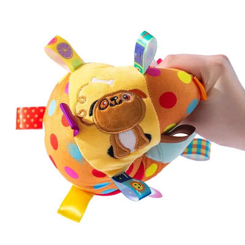 Balón interactivo de peluche color amarillo brillante y naranja con lunares coloridos, una cara de pug bordada en marrón y etiquetas de tela adheridas en diversos patrones con un asa.