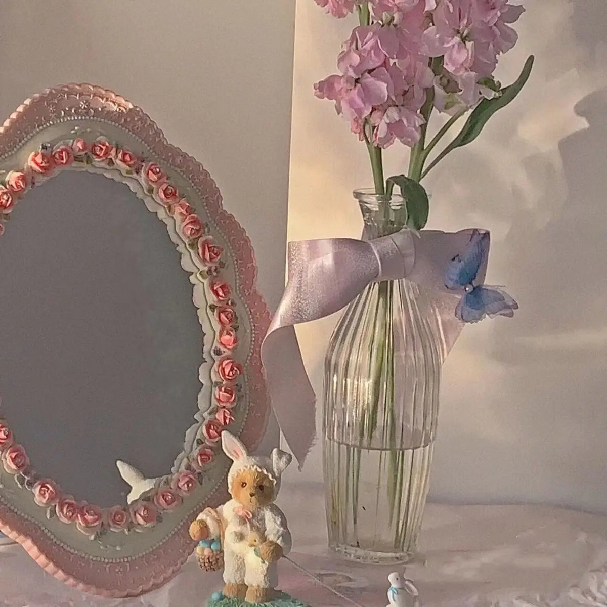Un jarrón de vidrio estriado transparente, con una cinta suave color lavanda y una decoración de mariposa azul colocada junto a un espejo de pared ovalado barroco color rosa con decoración floral vintage.