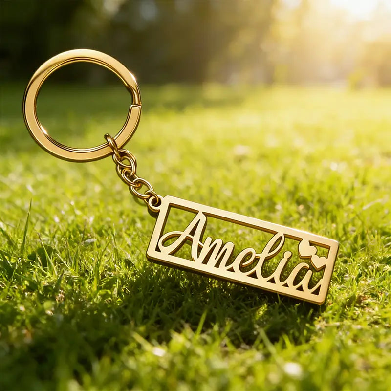 Gold-colored stainless steel custom name keychain featuring the name amelia engraved in cursive with small heart details attached to a circular ring by a short chain.