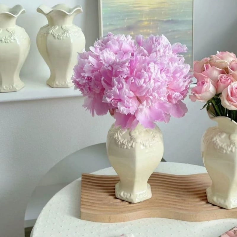 Un jarrón de cerámica moderno francés color crema con intrincados detalles de relieve floral que sostiene un exuberante ramo de suaves peonías rosadas como decoración elegante del hogar.
