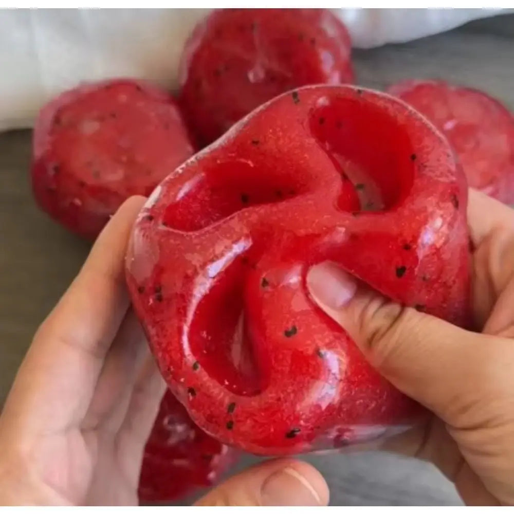 una bola antiestrés de rebote lento con forma de fruta del dragón roja brillante, con motitas tipo semillas negras y una textura esponjosa translúcida, diseñada como juguete artesanal de granizado de sandía para aliviar el estrés