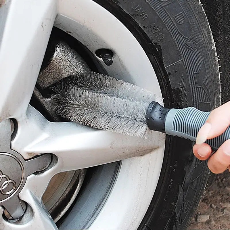 Une brosse de nettoyage pour roues de voiture en microfibre à soies grises, avec une poignée texturée noire et grise, est utilisée pour nettoyer les rayons d’une jante en alliage argentée.