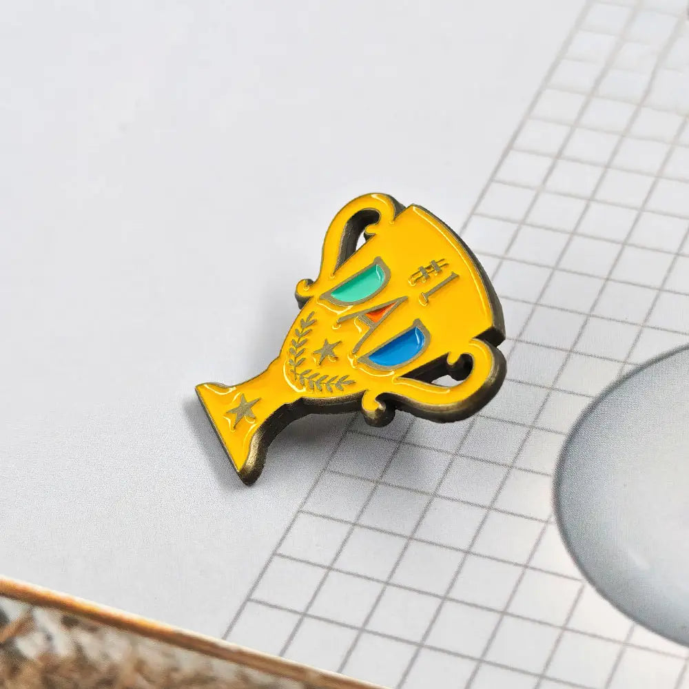 Broche en émail jaune éclatant en forme de trophée stylisé avec des yeux turquoise et bleus, des accents d’étoiles dorées, et le chiffre 1 sur son visage, indiqué comme broche dorée en émail de trophée « #1 Dad ».