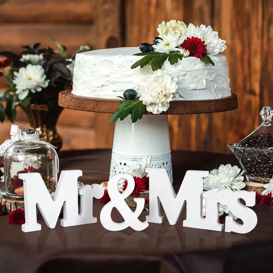 Ein weißer, mit Frost belegter Hochzeitstorte mit einer strukturierten Oberfläche, verziert mit frischen roten und weißen Blumen sowie Blaubeeren, neben einem hölzernen Hochzeitstischschild mit der Aufschrift „Mr“ und „Mrs“ dekoriert.