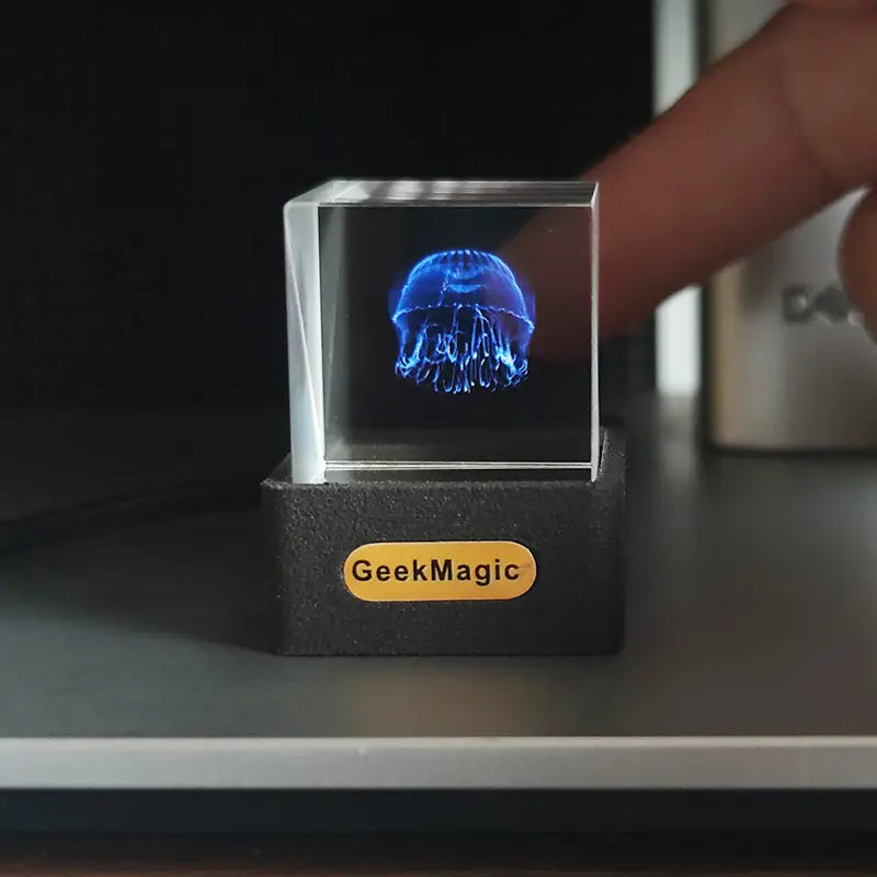 A glowing blue jellyfish hologram displayed inside a transparent acrylic cube on a black base with the Geekmagic logo as part of the Geekmagic smart weather station crystal gif clock.