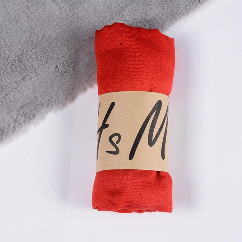 Ein leuchtend roter Baumwoll-Winterschal für Damen mit weicher Textur, ordentlich eingerollt und umwickelt mit einem beigen Papierband mit schwarzer, kursiver Beschriftung.