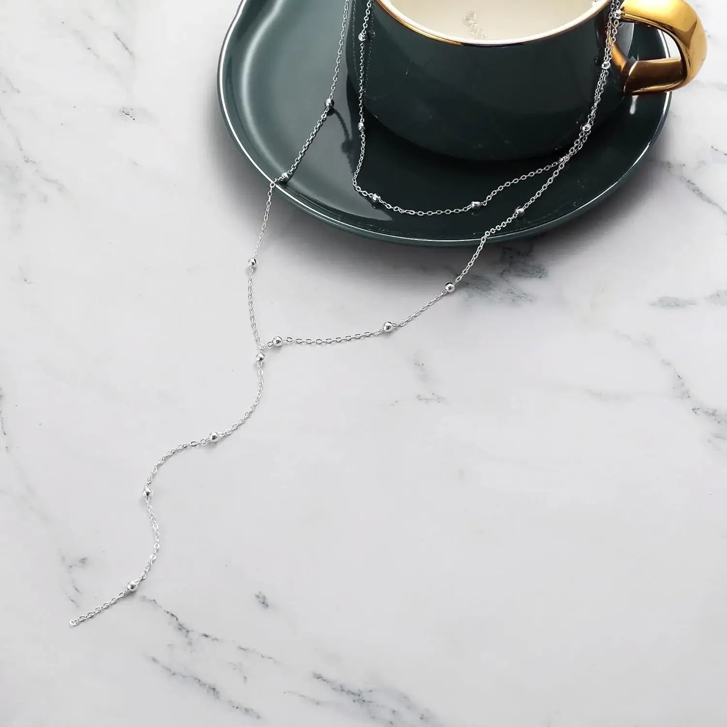 Un collier en chaîne à couches rehaussé de perles en argent, élégamment drapé sur une coupe et sa soucoupe en céramique vert foncé, dans un collier en couches d’or en forme de y pour produit femmes.