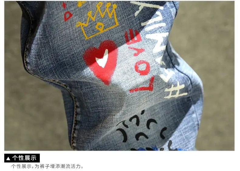 Jean slim imprimé stretch en denim bleu pour homme avec des graphismes colorés de style graffiti, incluant un cœur rouge, une couronne jaune et un texte blanc, avec « love » et « family ».