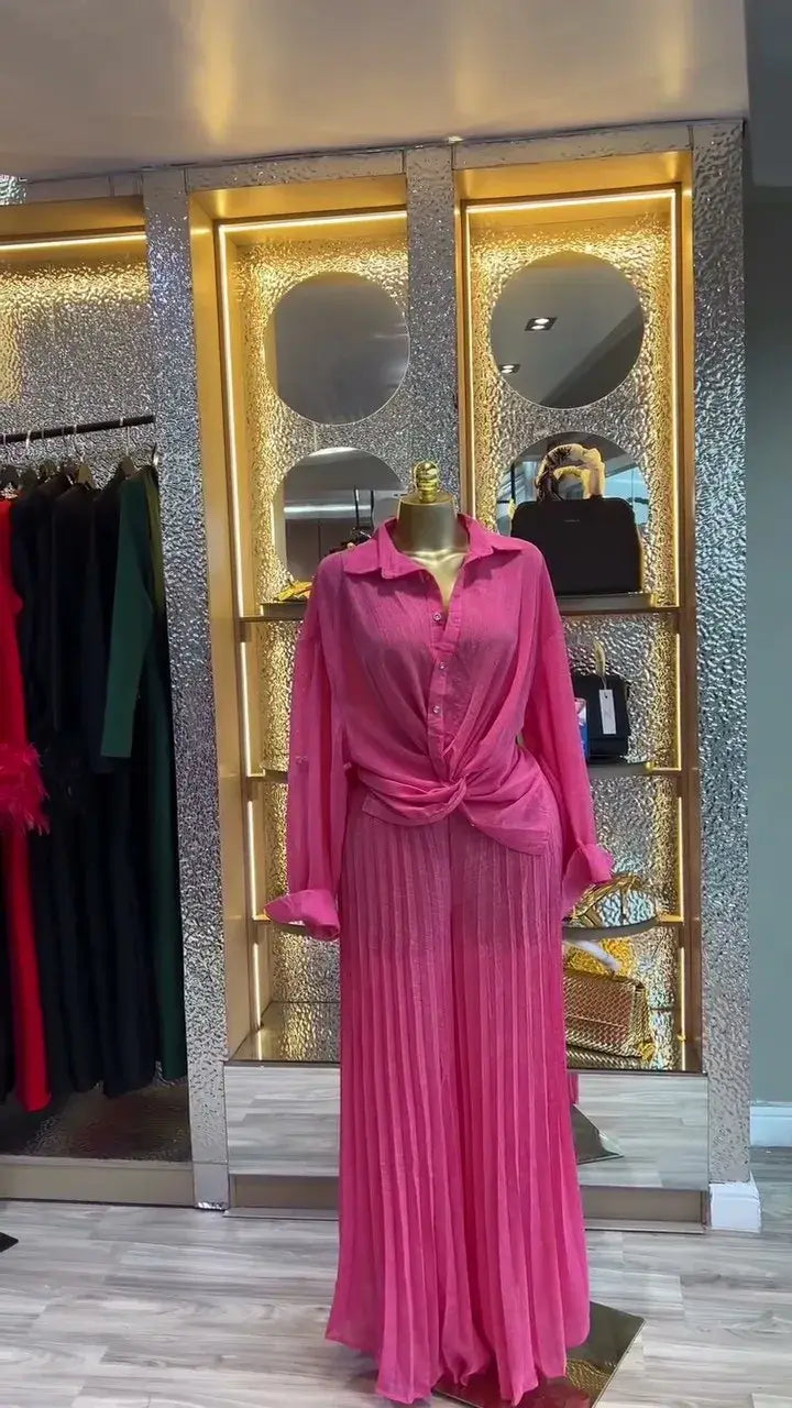 Ein lebhaftes pinkfarbenes Zweiteiler-Outfit mit langärmeliger Knopfbluse und passendem Falten-Maxirock, auf einem Schaufensterkörper präsentiert für das Damen-Set mit Faltenrockhose im weitem Bein, bestehend aus einem langärmeligen Outfit.