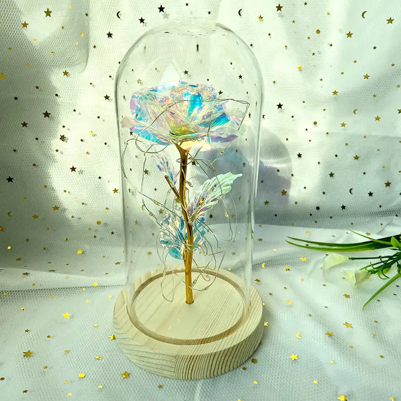 Eine irisierende blaue und violette Rose mit goldenem Stiel, umschlossen in einer klaren Glaskuppel auf einem hellen Holzsockel, im „Eternal Gold Rose Glass Dome Led Light Gift“.
