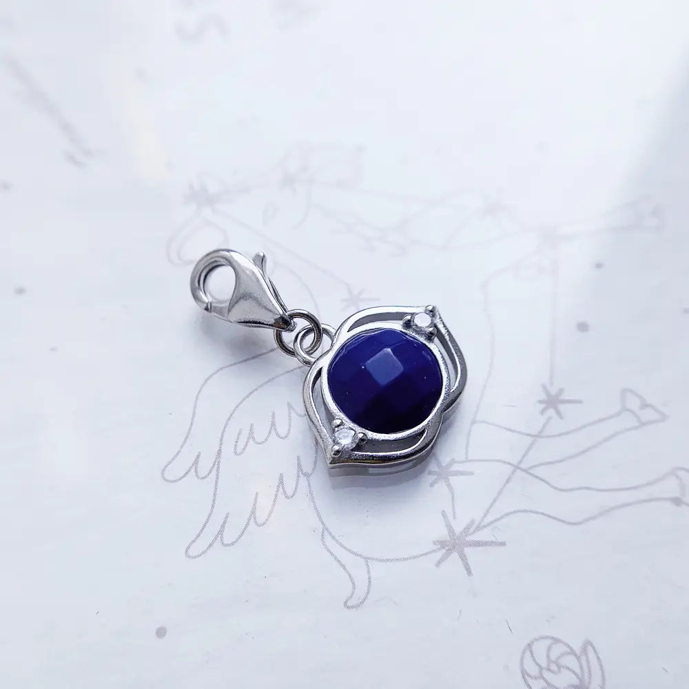 Colgante de encanto de plata de ley con acabado tono plateado que presenta una gema facetada azul profundo engastada en un marco ornamentado con forma de ala, realzado con dos pequeñas piedras transparentes, titulado Flor de Loto Azul.