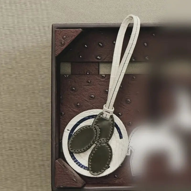 Une étiquette de bagage en cuir végétal vert olive, avec une lanière blanche gaufrée façon crocodile et une base à effet marbre circulaire, utilisée comme charm de sac à main de créateur.