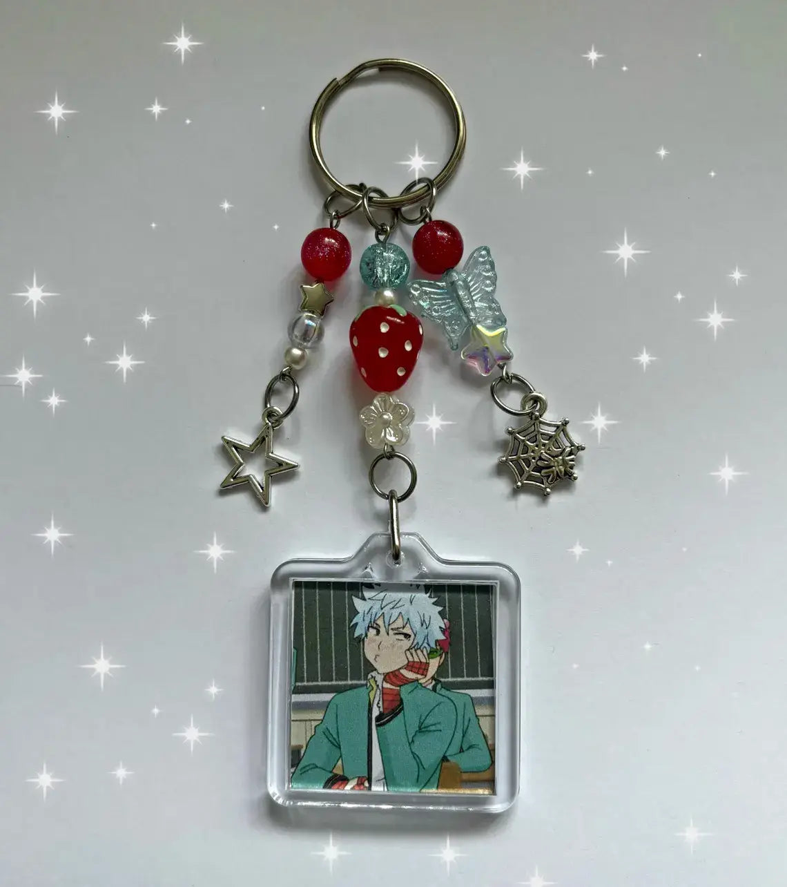 Clear acrylic Saiki k beaded anime keychain featuring a silver haired anime character in a green jacket decorated with red beads, a strawberry charm, and star charms.