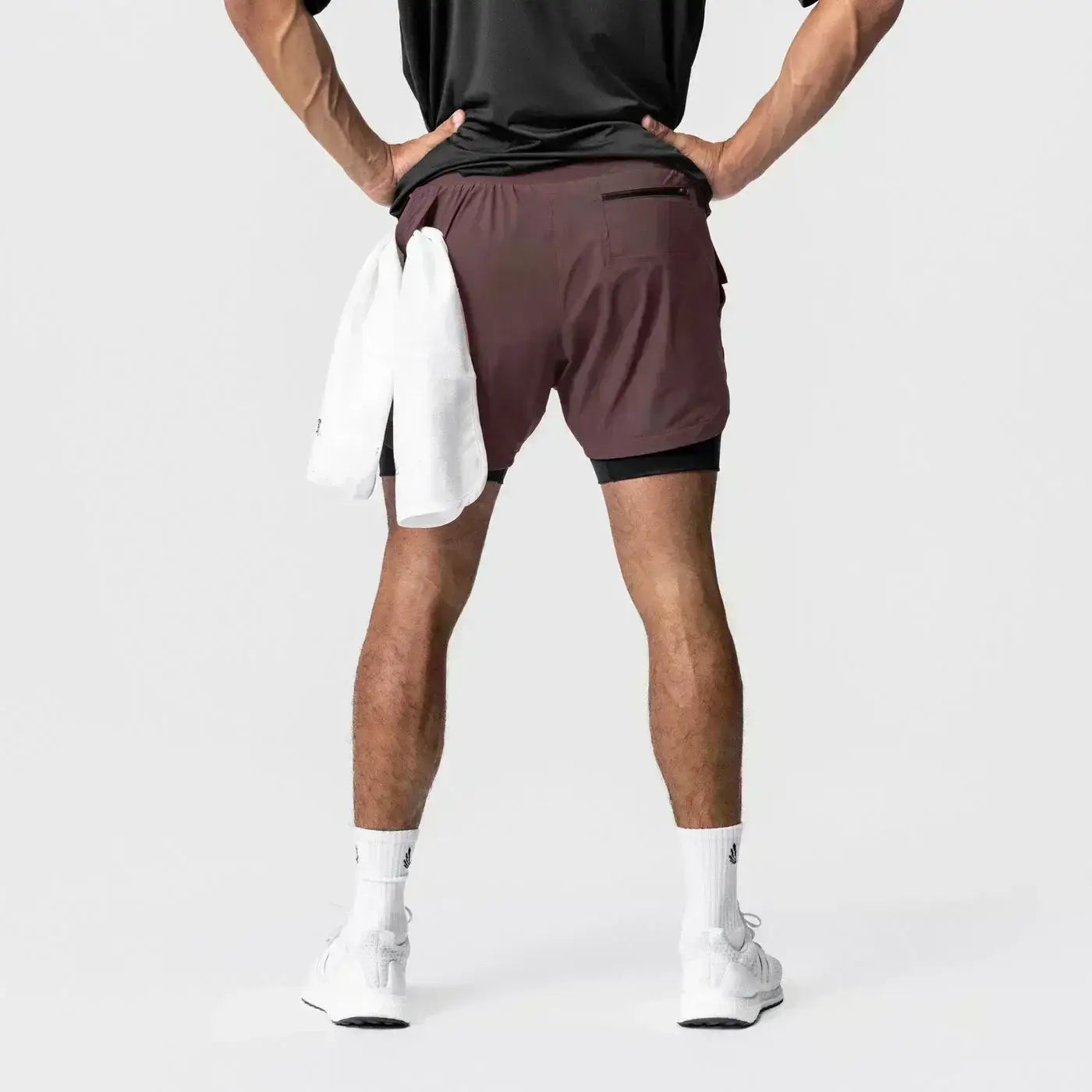 Hombres, pantalones cortos atléticos 2 en 1 para gimnasio en color marrón con capa interior negra, cintura elástica, bolsillo lateral y toalla blanca colocada sobre la parte posterior, con trabilla para la toalla.