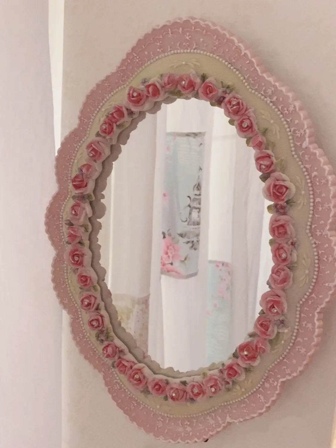 Un delicado espejo de pared ovalado barroco color rosa, con un marco festoneado adornado con adornos de rosas elaboradas a mano y acentos perlados para una decoración floral vintage.