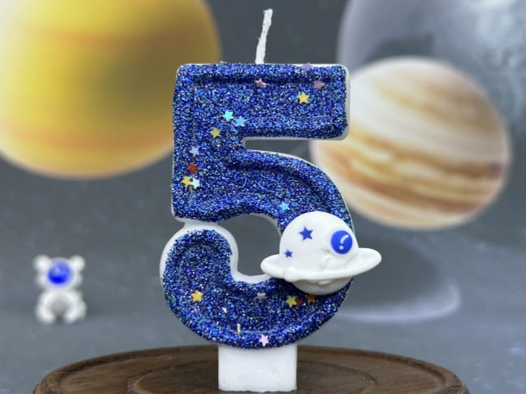 Una vela azul con brillo con el número cinco para cumpleaños, con un dije de nave espacial blanco que presenta detalles de estrellas y un solo ojo, utilizada como adorno para pastel de fiesta planetaria.