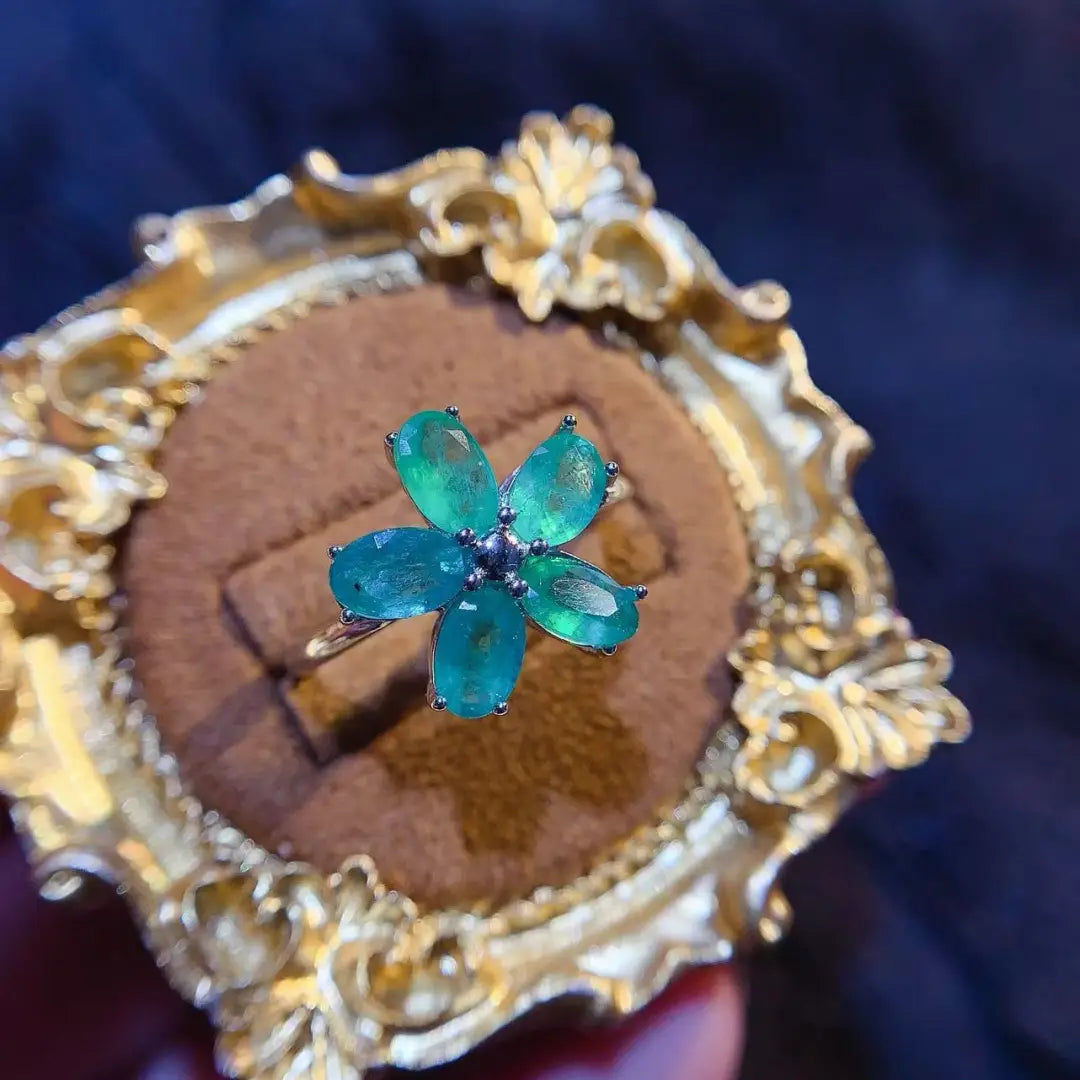Ein zarter, blütenförmiger Smaragdblumen-Silberring mit fünf leuchtenden emeraldgrünen Edelsteinen, die in einem silberfarben wirkenden Metallband eingefasst sind.