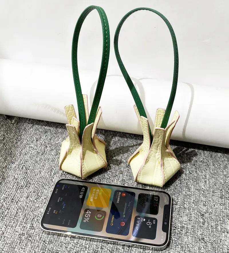 Un duo de charms pour sac à main en cuir végétal gaufré façon crocodile, couleur crème, élégants, avec des poignées vertes fines, conçus dans une forme unique évoquant une tulipe.