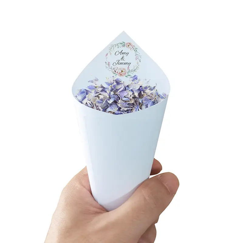 Eine weiße Papiertüte, gefüllt mit lila- und weißen getrockneten Blütenblättern, mit einem personalisierten floralen Kranzdesign an der Oberseite, das „amy“ und „jimmy“ liest, im Set von 30 personalisierten Hochzeits-Konfetti-Tüten.