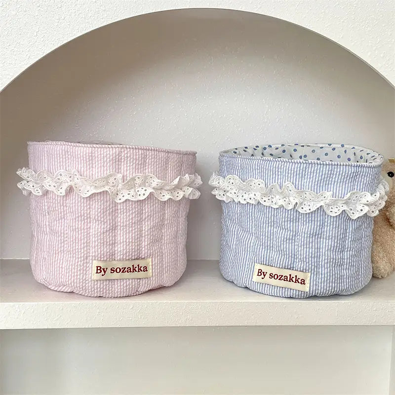 Gepolsterte Aufbewahrungskörbe aus pastellrosa und hellblau gestreiftem Stoff mit weißem Spitzenbesatz zur geordneten Aufbewahrung im Babyzimmer
